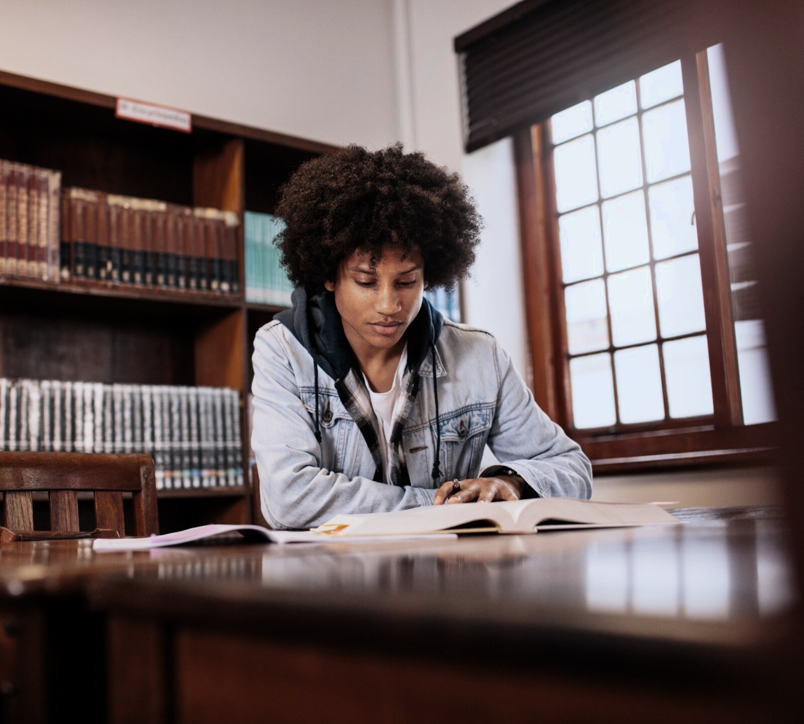 Young man studying an open book at a wooden library table.